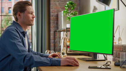 Fototapeta na wymiar Young Handsome Man Working from Home on Desktop Computer with Green Screen Mock Up Display. Male Checking Corporate Accounts, Messaging Colleagues. Loft Living Room with Big Window.