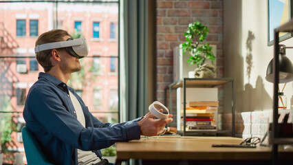 Portrait of a Man Sitting at a Desk at Home or Creative Agency, Using a Virtual Reality Headset and Controllers. Young Male Working from Living Room, Experiencing Next Generation of Internet Services.