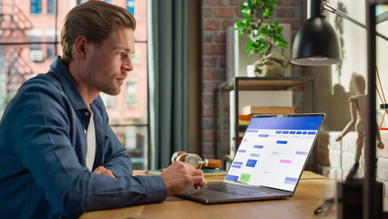 Young Handsome Man Working on Freelance from Home on Laptop Computer with Office Software on Display. Creative Male Checking Timetable, Planning Meetings. Living Room in Loft City Apartment.