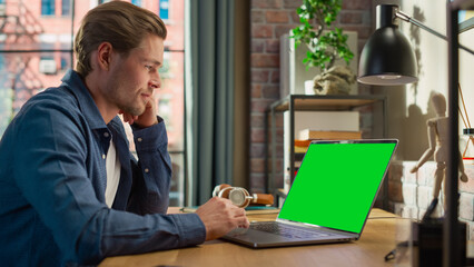 Young Handsome Man Working from Home on Laptop Computer with Green Screen Mock Up Display. Creative...