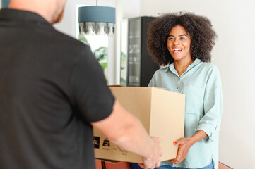 Back view of courier giving cardboard box to happy african american woman. Cheerful multiracial girl with natural Afro hairstyle receiving a package at home from a delivery guy, ordered online - Powered by Adobe
