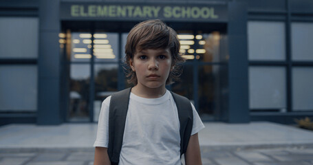 Sad elementary age boy near modern school entrance. Teen student looking camera.