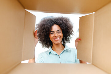 POV view from inside box at satisfied biracial young woman with an Afro hairstyle unpacking package box, excited happy African woman looks inside delivery box. Glad customer of e-shop - Powered by Adobe