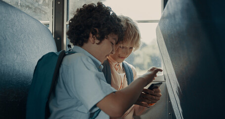 Two pupils playing games on phone sitting school bus. Boys using smartphone.
