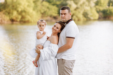 Happy family day. Mother, father and baby daughter spending free time outdoors near lake, pond. Family enjoying time together at meadow. People having fun in nature. Family bonds outside.