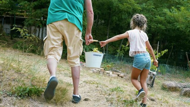 Grandfather and granddaughter carrying organic waste bucket with food leftovers into composter