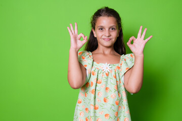 Portrait image of very happy amazed smiling brunette little cute girl, in summer dress, showing okay or zero hand sign gesture, isolated over green background with copy space.