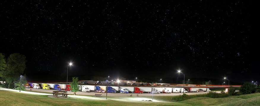 Trucks in rest area at night. Truck stop wayside, parked. Wide view with copy space and lighted foreground. Stars in the sky, background.