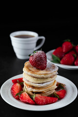 On a dark background, pancakes with powdered sugar and strawberries in a white plate, in the background a cup of coffee and strawberries