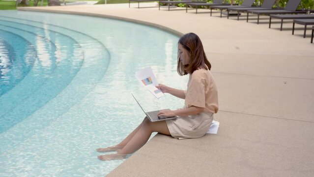 4K, medium shot A woman working at the swimming pool with her laptop computer holding paperwork in her hand. It is imperative for young Asian female tourists to sit and work on vacations at the hotel.