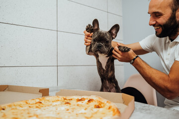 Cheerful Man Holding French Bulldog by Paws in Front of Pizza on the Kitchen Desk, Small Dog Impatiently Waiting for Human Food