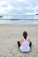 Fototapeta na wymiar Young woman wearing white blouse sitting alone on the sand at Ribeiro Beach, Bombinhas, Santa Catarina state, Brazil