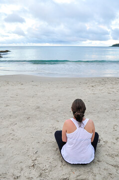 Young woman wearing white blouse sitting alone on the sand at Ribeiro Beach, Bombinhas, Santa Catarina state, Brazil