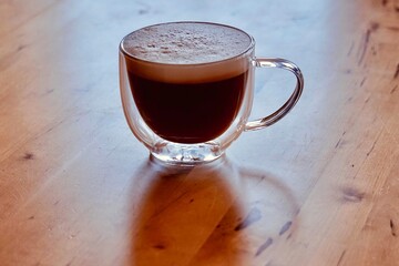Kaffeetasse aus Glas gefüllt mit Milchschaum Spielgelung auf der Tischplatte aus Holz