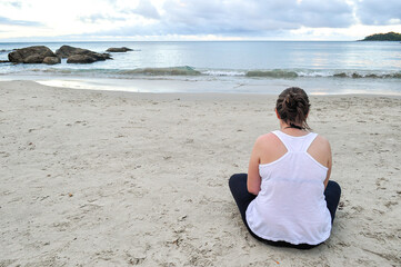 Fototapeta na wymiar Young woman wearing white blouse sitting alone on the sand at Ribeiro Beach, Bombinhas, Santa Catarina state, Brazil