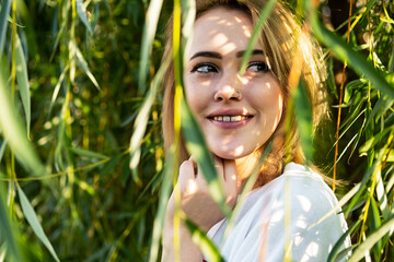 Fototapeta premium Cute woman smiling under willow tree in sunlight with shadow from branches. Female portrait in woodland on sunny day.
