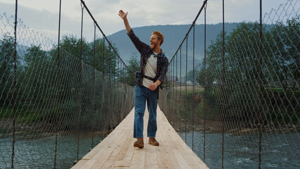 Fototapeta na wymiar Smiling traveler waving hand on river bridge. Hiker enjoy holiday in mountains.