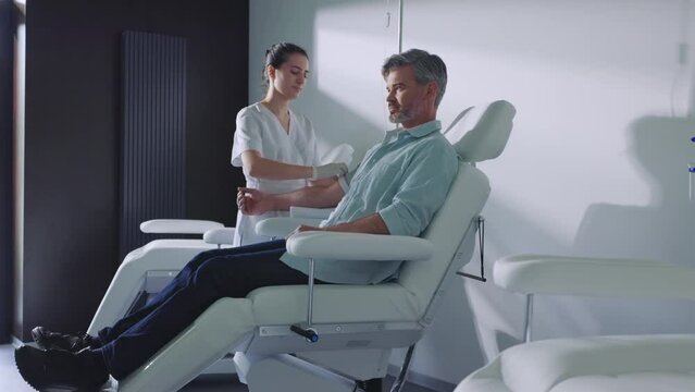 In a modern clinic doctor finishes taking blood from the patient in light room. Female nurse and patient man sitting talking smiling. Slow motion