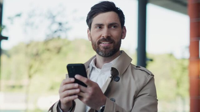 Smiling young stylish handsome man with beard in coat uses smartphone stand in part at sunlight. Close up. Slow motion