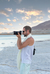 Adult cheerful man photographer taking photographs with digital camera. Travel, vacations, work and active lifestyle concept. Handsome muscular man using picture with modern dslr camera at the beach