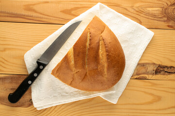 One half of a fragrant fresh loaf with a white linen napkin and a knife on a wooden table, close-up, top view.