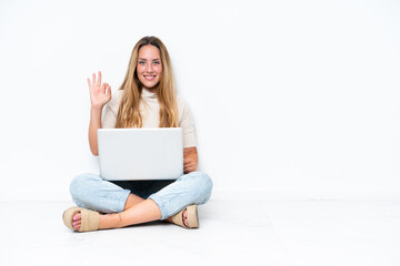 Young woman with laptop sitting on the floor isolated on white background showing ok sign with fingers