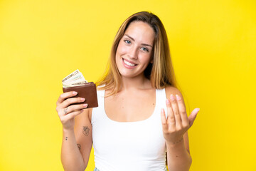 Young caucasian woman holding a wallet isolated on yellow background inviting to come with hand. Happy that you came