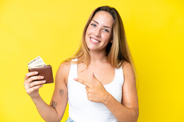 Young caucasian woman holding a wallet isolated on yellow background and pointing it