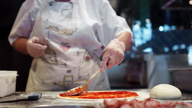 Chef in white clothes preparing pizza, spreading red sauce for pizza spreading dough with metal spoon, process of preparing dough with toppings for delicious Italian pizza in a street restaurant
