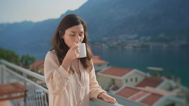 Young inspired asian woman standing terrace, enjoying drinking hot coffee, tea, watching views of sunrise landscape and scenery. People at home. Peaceful morning old European city in mountains by sea.