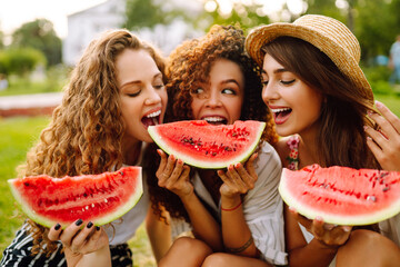 Three young woman  camping on the grass, eating watermelon, laughing. People, lifestyle, travel, nature and vacations concept.