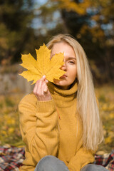 Attractive blonde woman in a yellow sweater is sitting on a plaid in an autumn park and covering half of her face with a yellow maple leaf.Autumn concept.Beauty in nature.