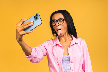 Young funny african american black casual woman grimacing and making selfie on smartphone isolated over yellow background.