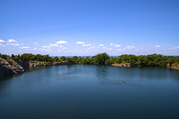 Scenic Landscape Of A Lake Surrounded By Trees, Cliffs, And Flat Rocks With The View Of Ocean In The Far Against A Partially Cloudy Blue Sky Under Which The Seagulls Enjoy The Clear Water