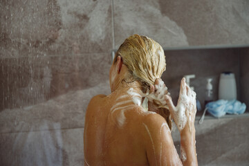 Rear on blonde Caucasian female washing hair in shower and cleaning with shampoo.