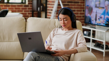 Attentive worker doing remote work on laptop while writing down important job informations. Focused woman taking notes while working remotely and listening to music at home in living room. Tripod shot