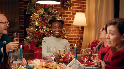 Woman at Christmas dinner table clinking glasses with family member while smiling at camera. People in living room celebrating winter traditional holiday with sparkling wine and home cooked food.