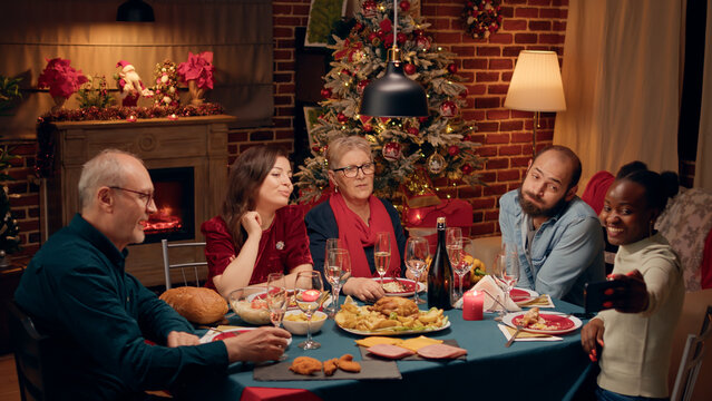 Happy festive multiethnic family taking selfie photo with modern smartphone while enjoying Christmas dinner at home. Joyful diverse people celebrating winter holiday together.