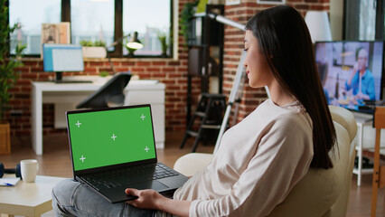 Young adult person sitting on couch while using laptop having chroma key isolated background. Woman sitting on couch inside apartment with portable computer having green screen display. - Powered by Adobe