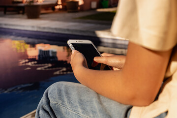 a teenager looks at her cell phone sitting by a pool. Unrecognizable person.
