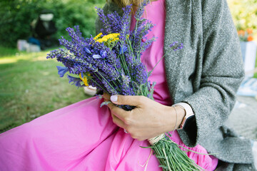 summer bouquet of yellow yarrow and lavender bouquet in woman's hands, woman wearing a pink dress sitting in a park - 521531973