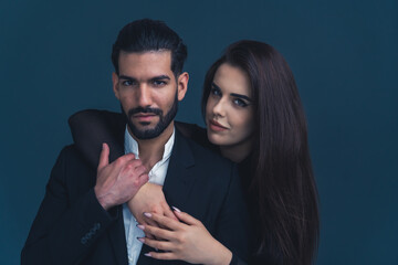White woman leaning seductively on her husband wearing formal clothes from the back putting hands on his chest. Dark background studio shot. High quality photo