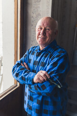Retired caucasian senior bald man with arms crossed, holding his glasses and smiling at the camera while standing near the window. High quality photo