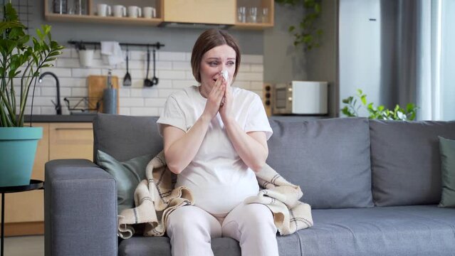 sick pregnant woman with cold cough and temperature blowing nose with runny nose sitting on sofa at home. ailing female got flu during pregnancy. Disease. suffering Allergy cold symptoms