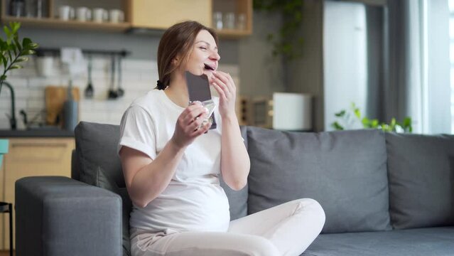 Hungry pregnant woman with belly eats sweets unhealthy and junk food. Female with appetite hold plate and eating Dark chocolate cookies Sitting at home on sofa. Indoor. Sweet cravings during pregnancy