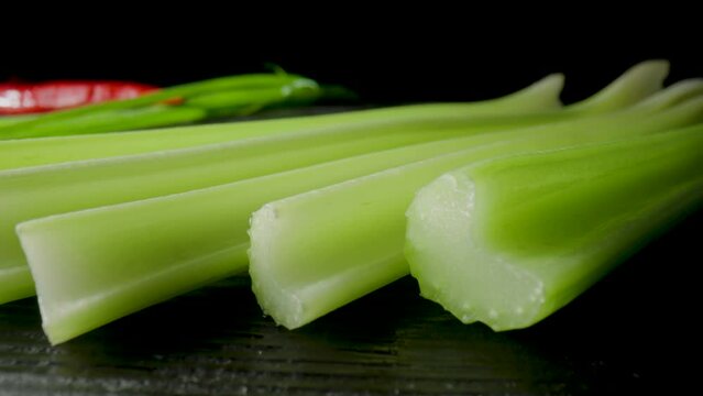 Row of green celery stalks lying on black board on isolated black studio background. Macro shot of fresh celery and green and red hot peppers. Ingredients for salad. Slow motion ready 59.94fps.