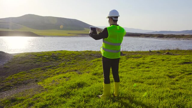 Engineer doing land and water survey.
The engineer, holding a tablet in the field, conducts investigations around the land and the lake.
