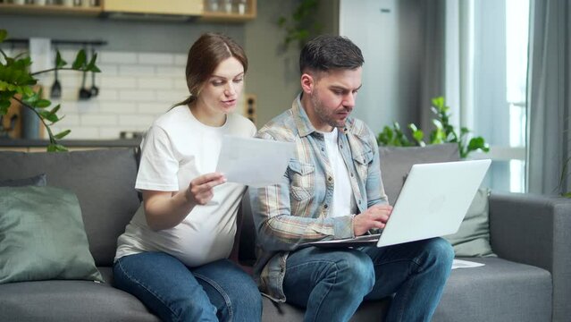 Young married couple, husband and pregnant wife together at home, satisfied sitting on sofa and working at laptop computer paperwork at home using website online easy and accessible to use