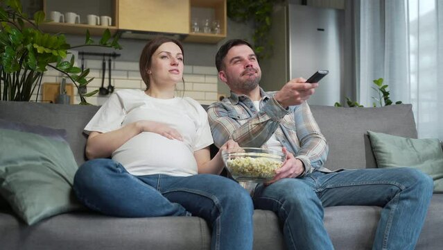 Bored couple at home choose a television show to watch and switch channels with a TV remote control. Young family husband and pregnant wife cannot choose a show. watching news or film during pregnancy