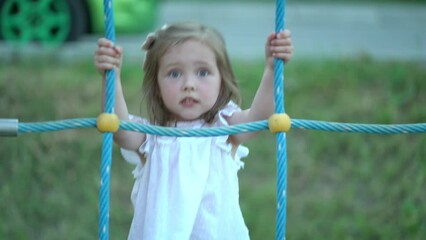 Three-year-old girl in a white dress on the playground. Cute little girl climbing on rope mesh at playground. Child climbing a rope ladder - Powered by Adobe